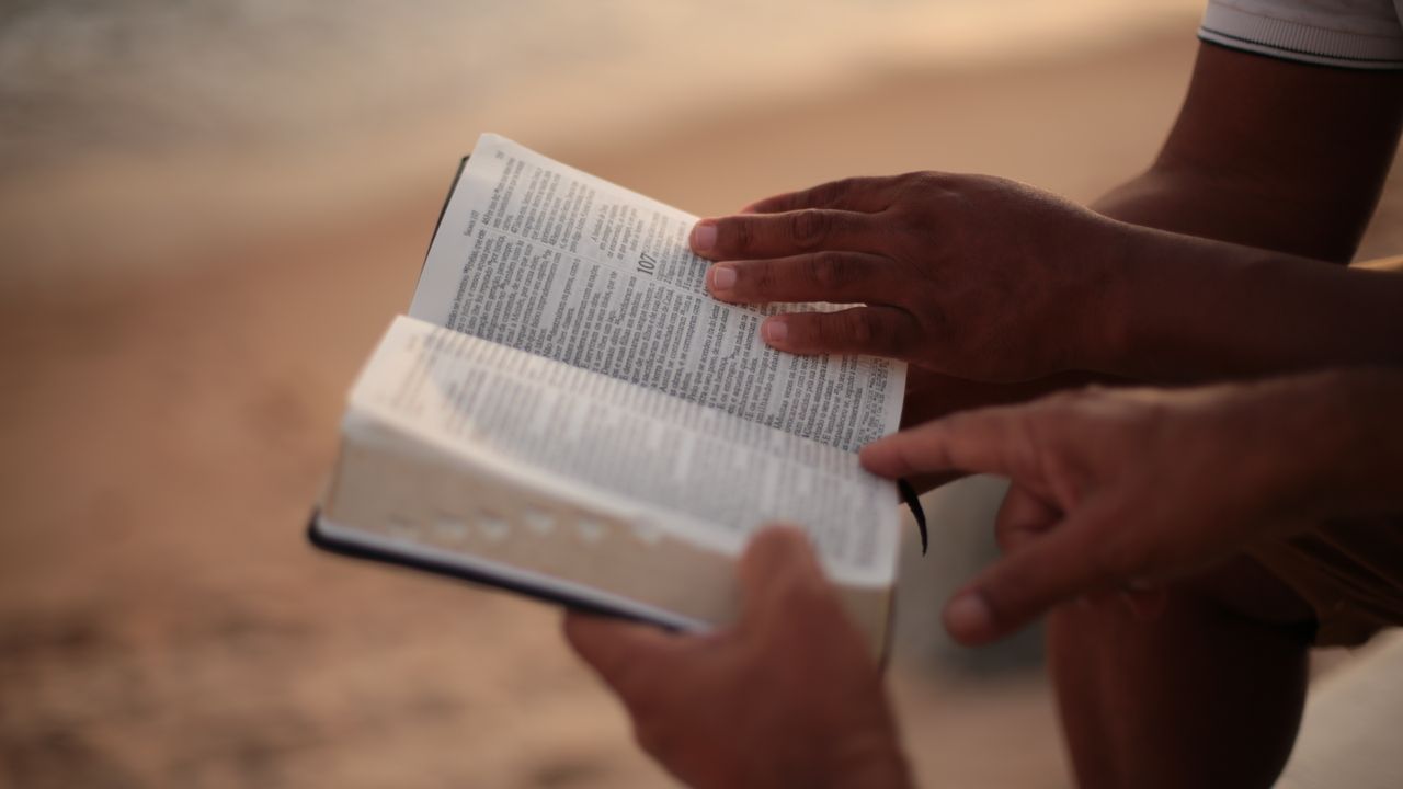 Services Close-up of hands holding an open Bible on a beach, symbolizing faith and study.